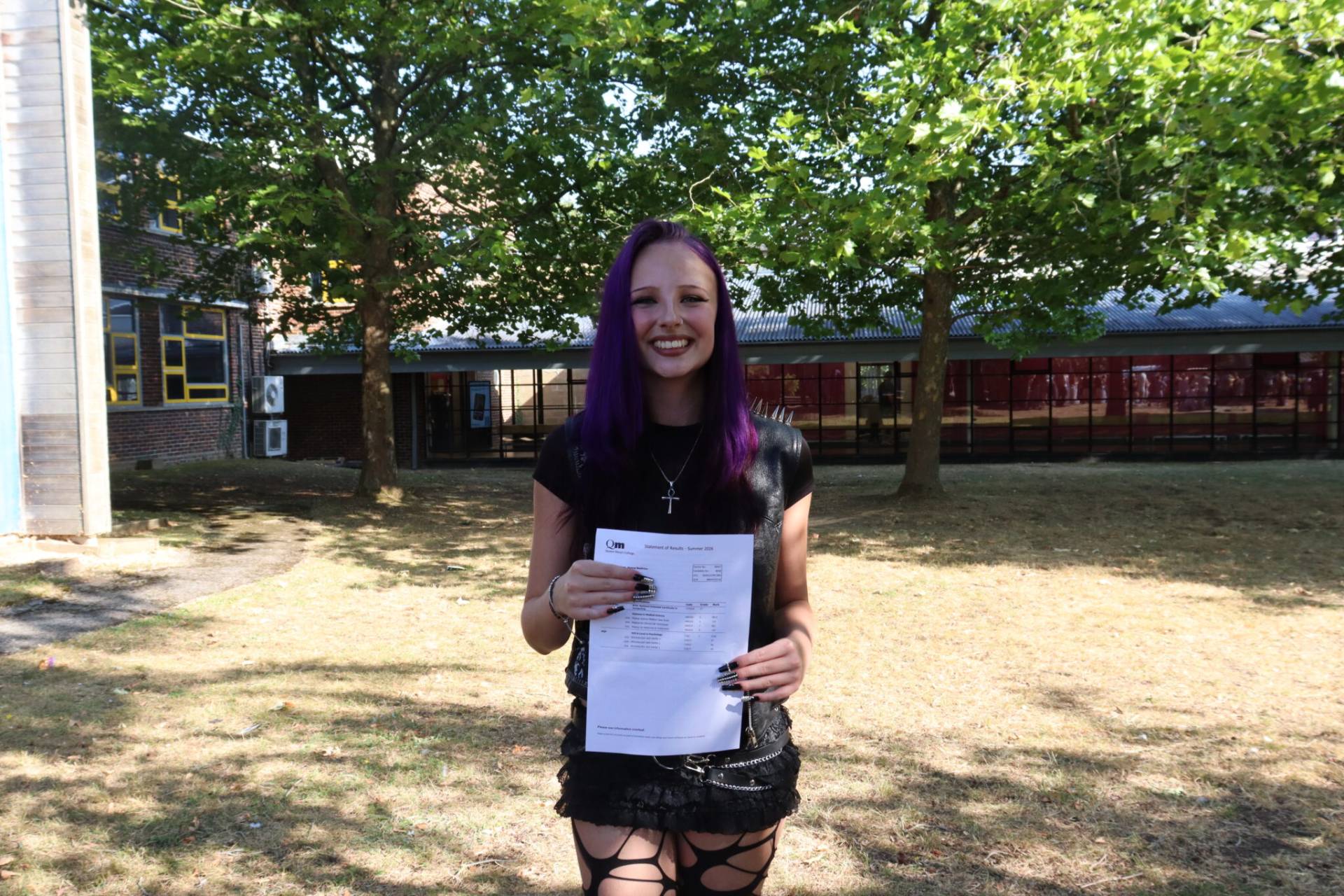Smiling young woman with purple hair holding a printed document outdoors on a sunny campus lawn, with trees and buildings in the background.