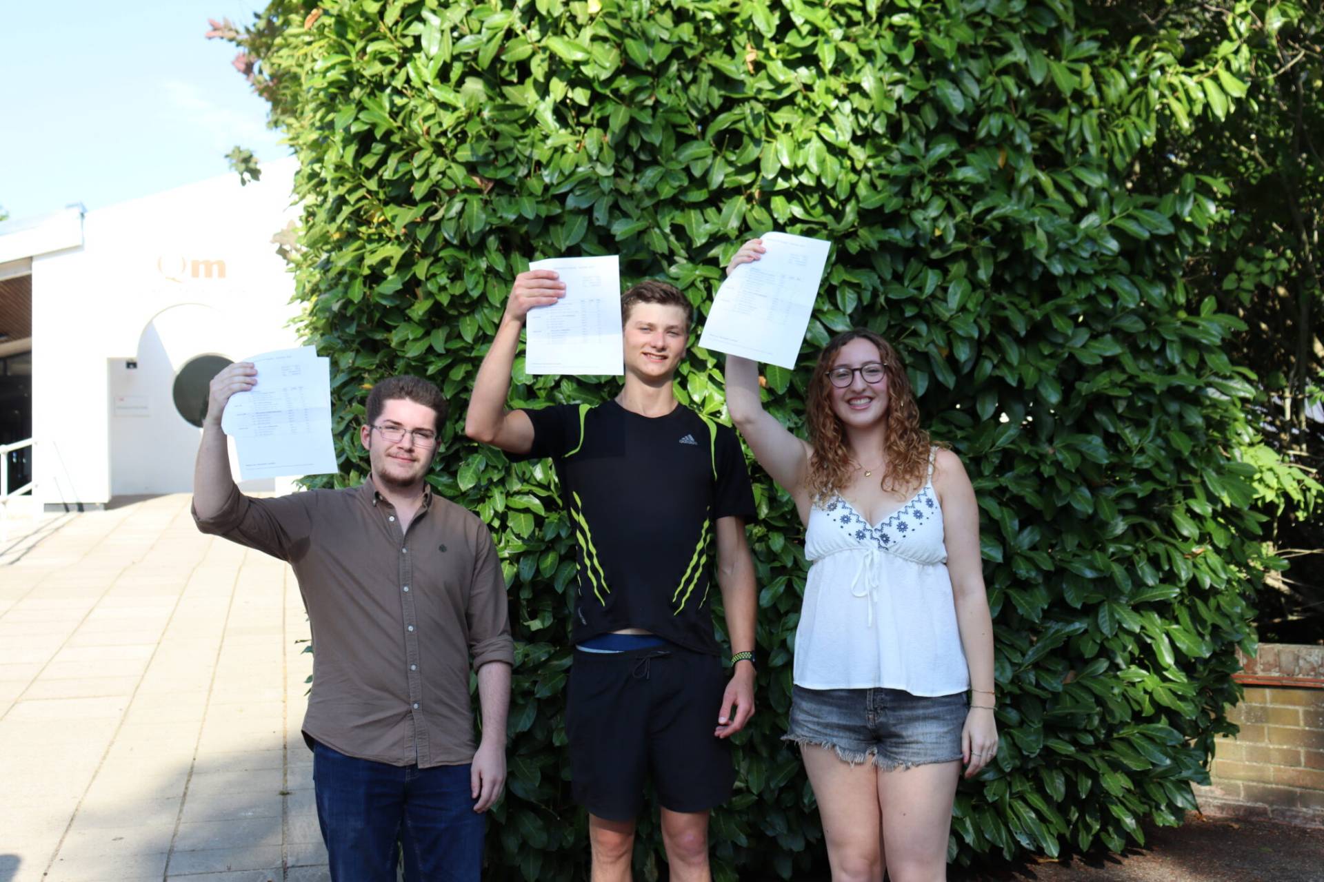 Three friends pose outdoors in front of a dense green hedge, each holding up printed documents and smiling at the camera.