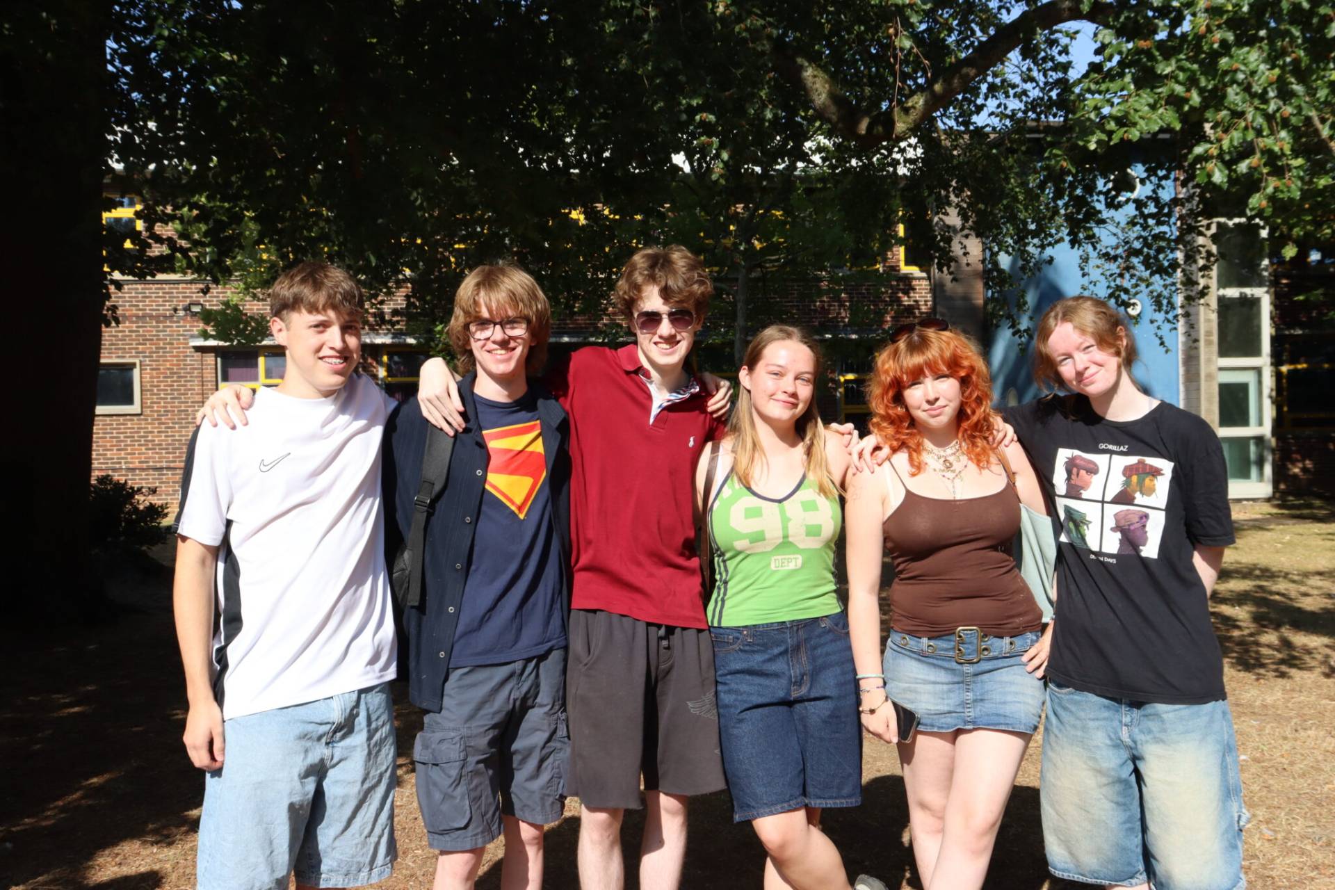 Six young people stand arm-in-arm outdoors, smiling, with leafy trees and a brick building in the background for a casual group photo.