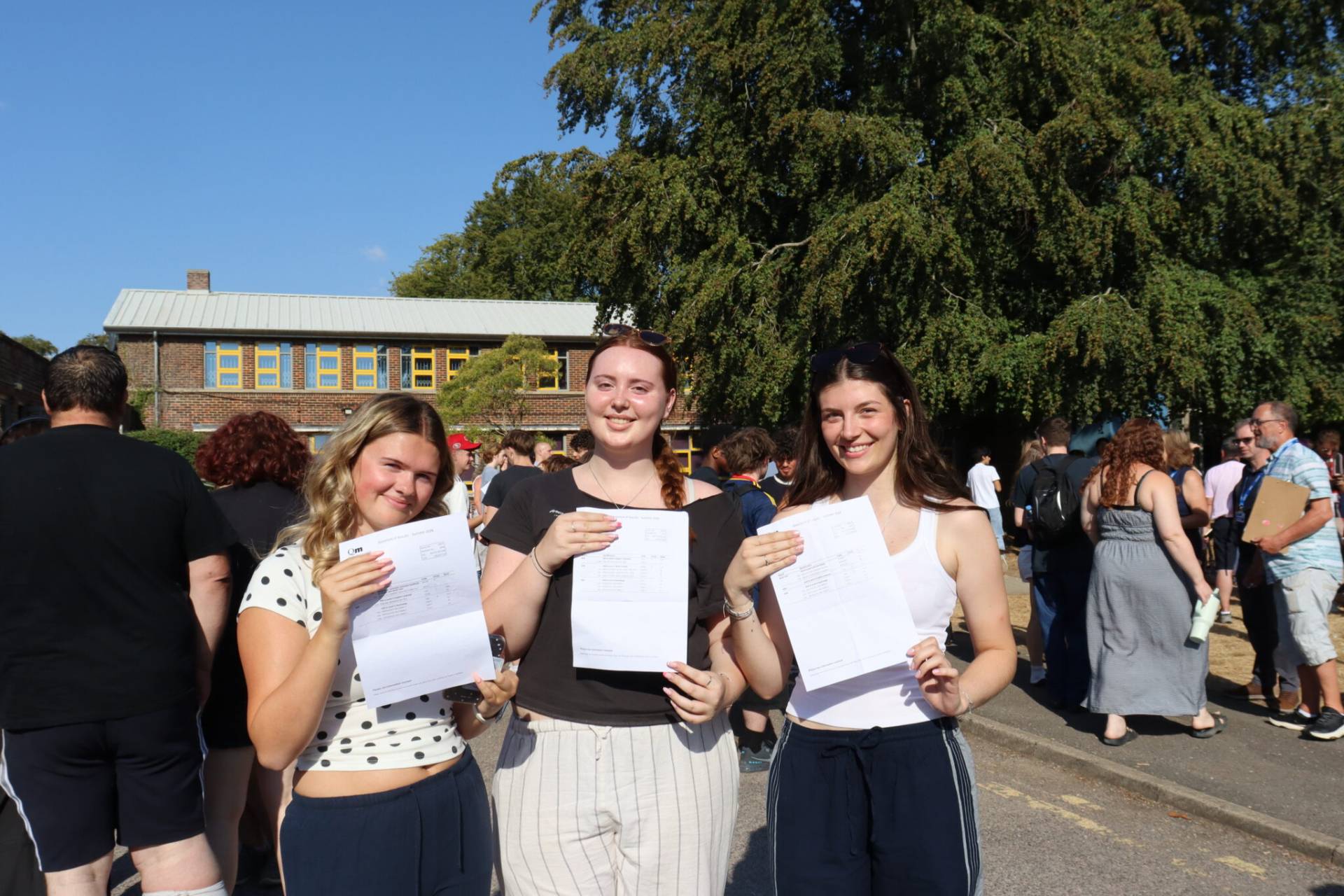 Three smiling students hold papers outdoors on a sunny day, with a brick building and large tree behind them.