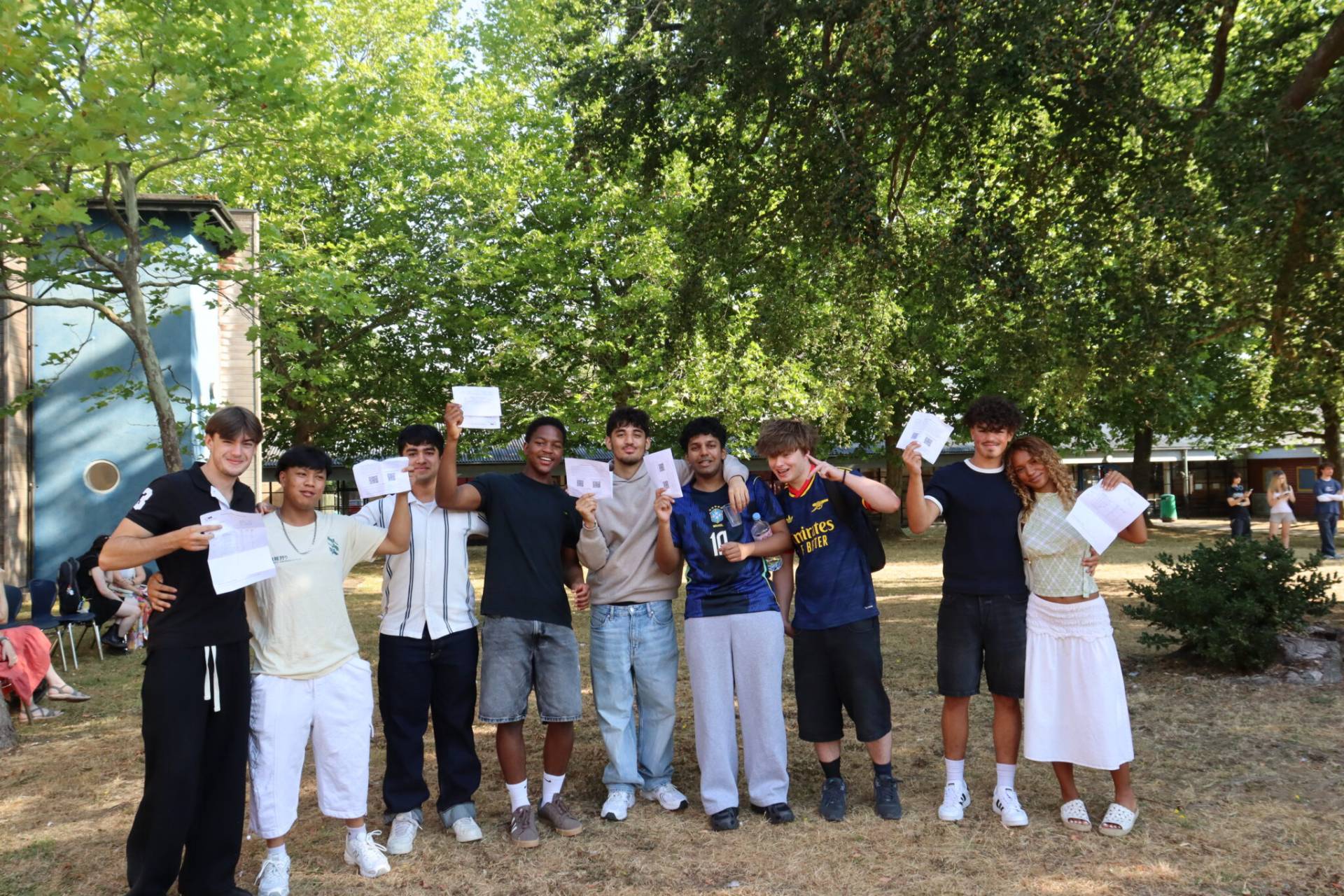 Group of teens outdoors, smiling and holding papers, with trees and a building in the background.