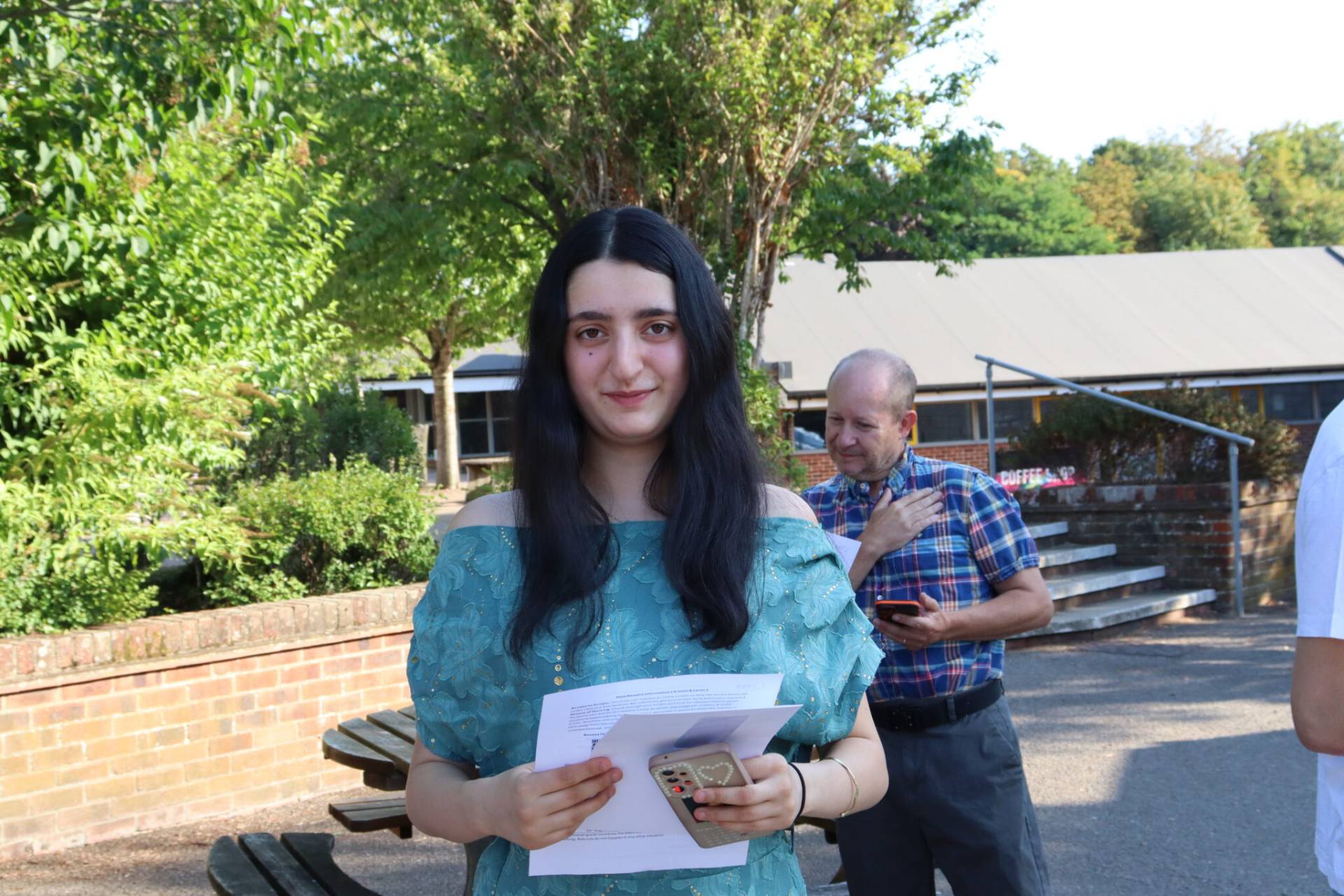 Young woman in a teal off-the-shoulder dress holds papers and a phone outdoors, with a man in a plaid shirt behind her.