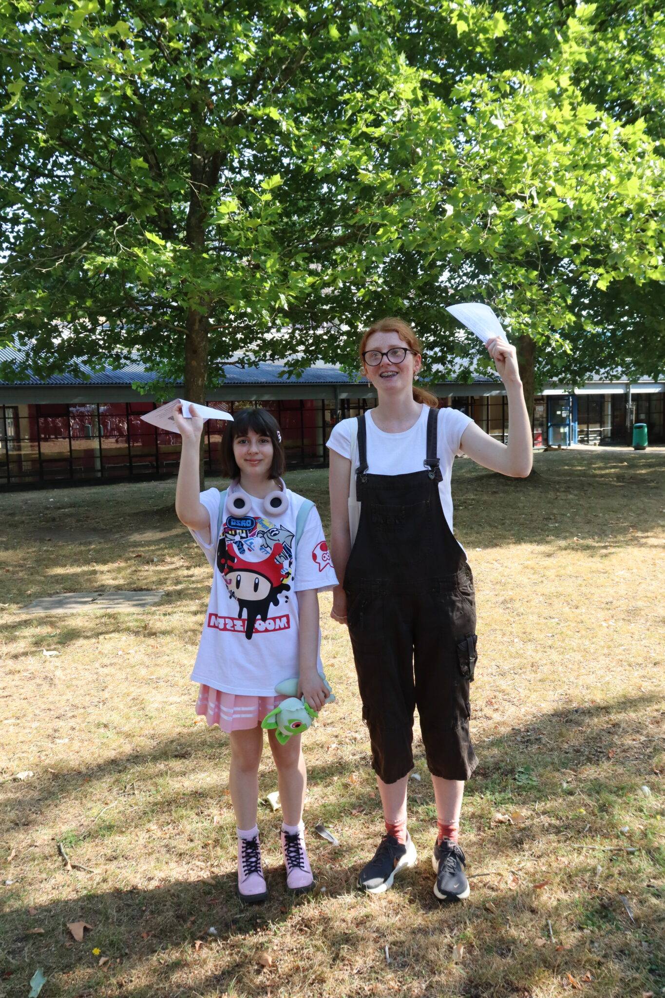 Two girls stand outdoors under leafy trees, holding papers as if showing something they made; a building is in the background.