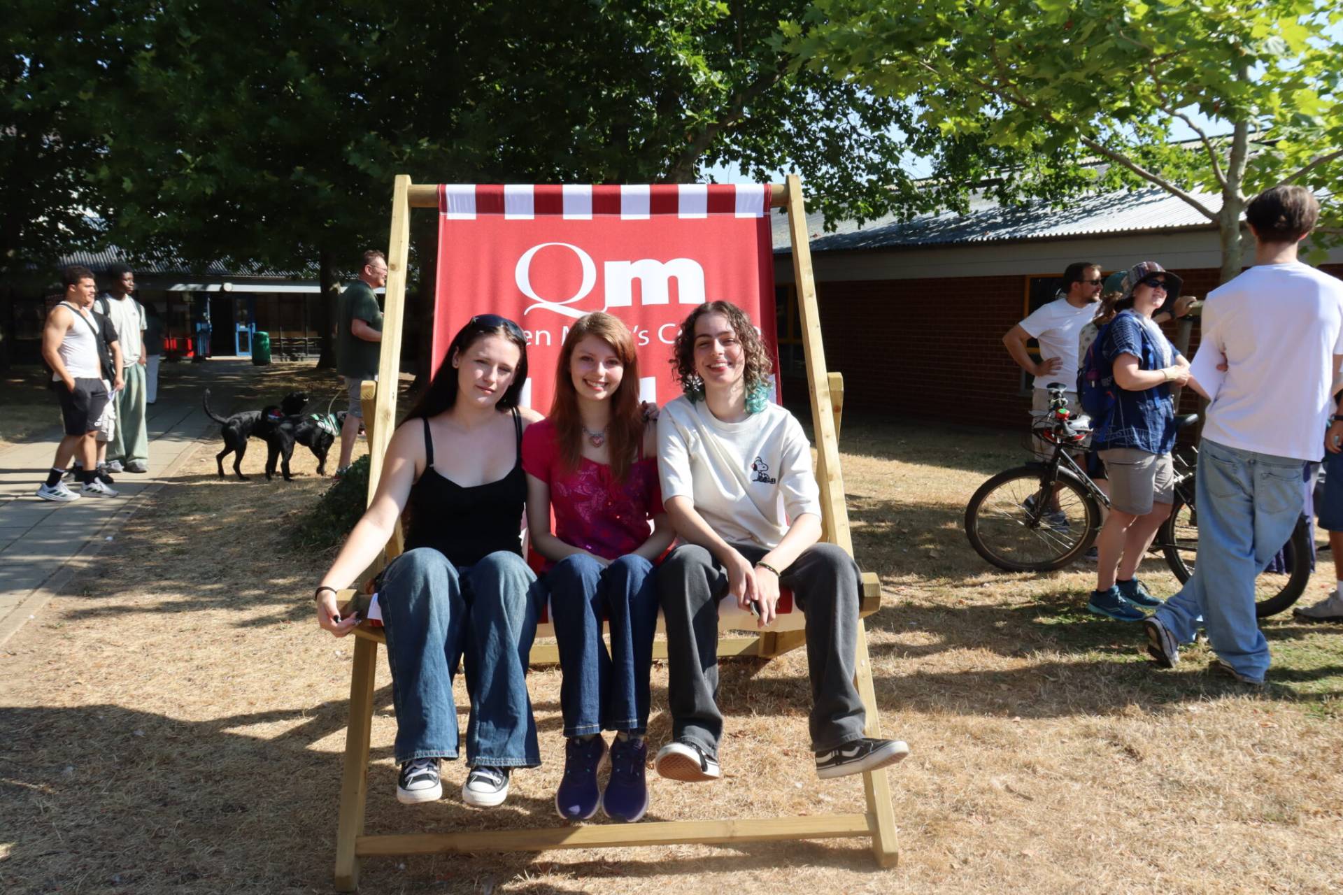 Three smiling young women pose on a large wooden chair outdoors, with a red banner behind them and people chatting in the background.