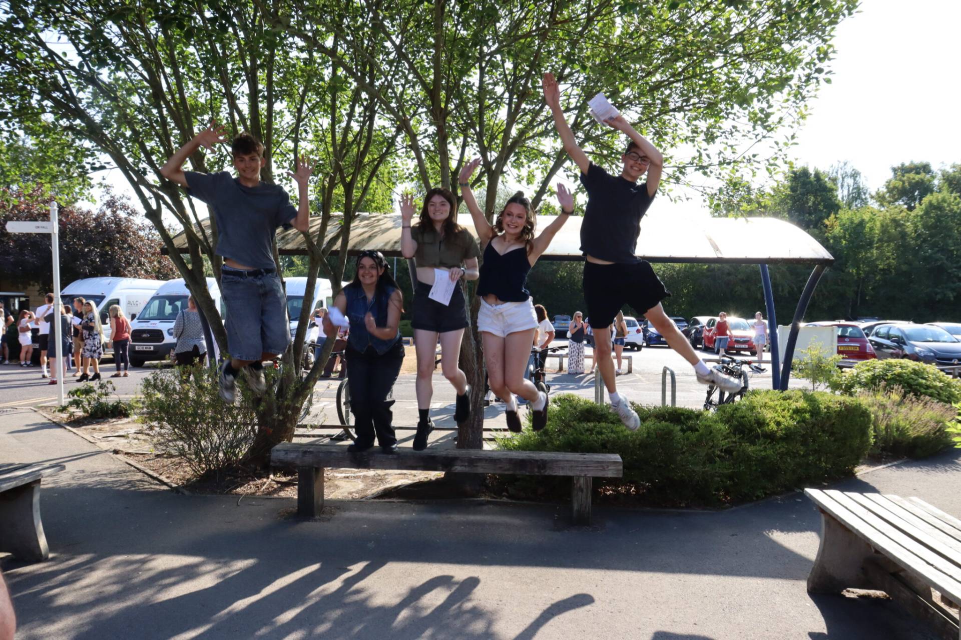 Five friends jumping mid-air off a wooden bench in a sunny outdoor area with trees behind them.