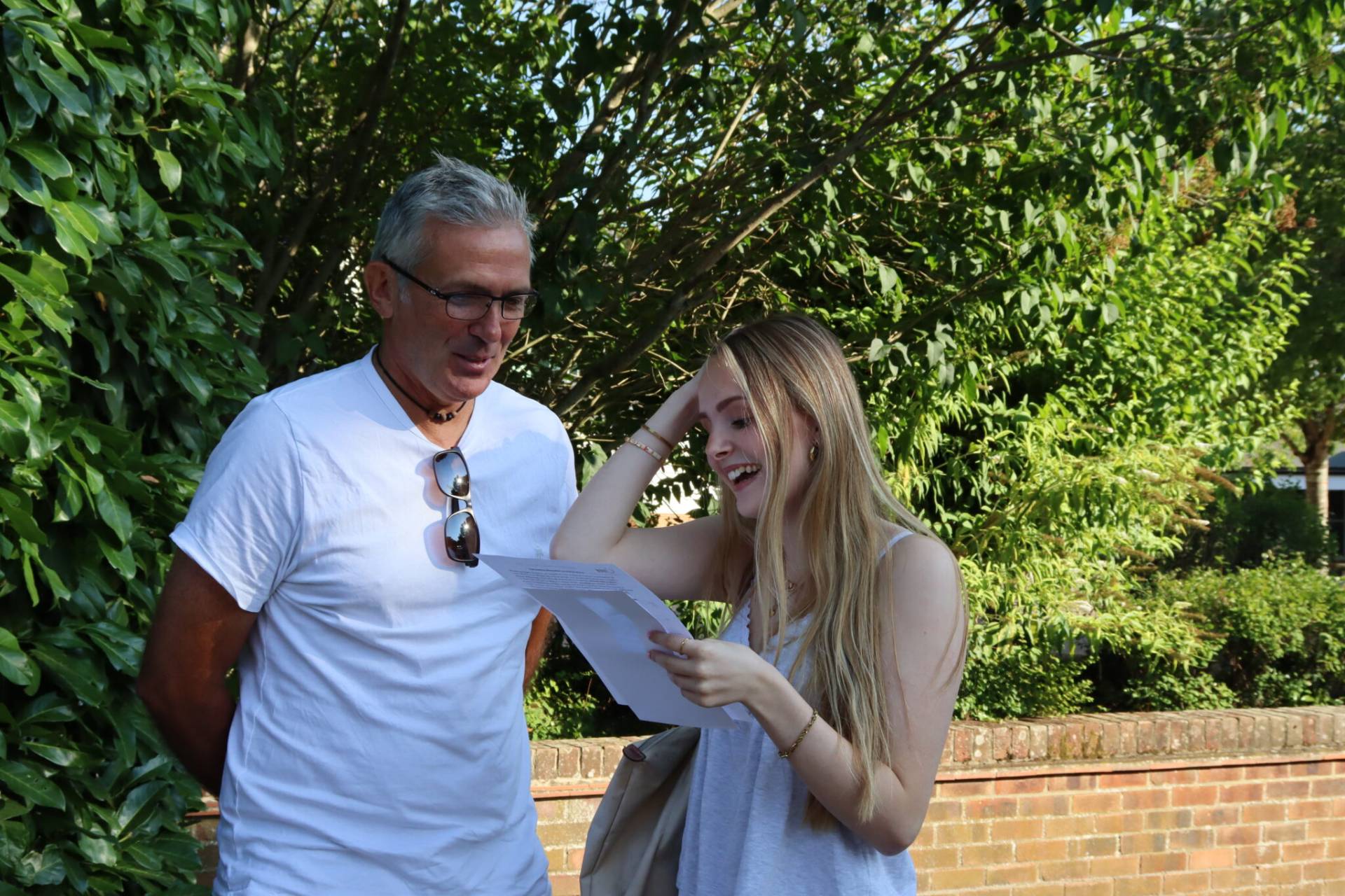 Man and woman outdoors examining a sheet of paper together, smiling as they read in front of greenery and a brick wall.