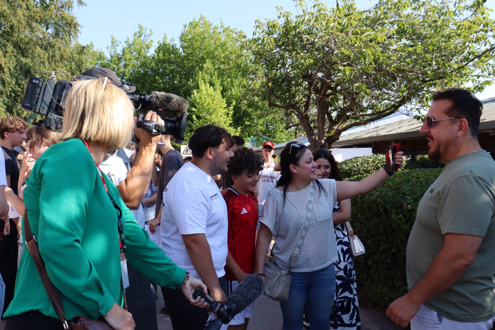 Cameraman filming a crowd of people outdoors with a large video camera and microphone under trees on a sunny day