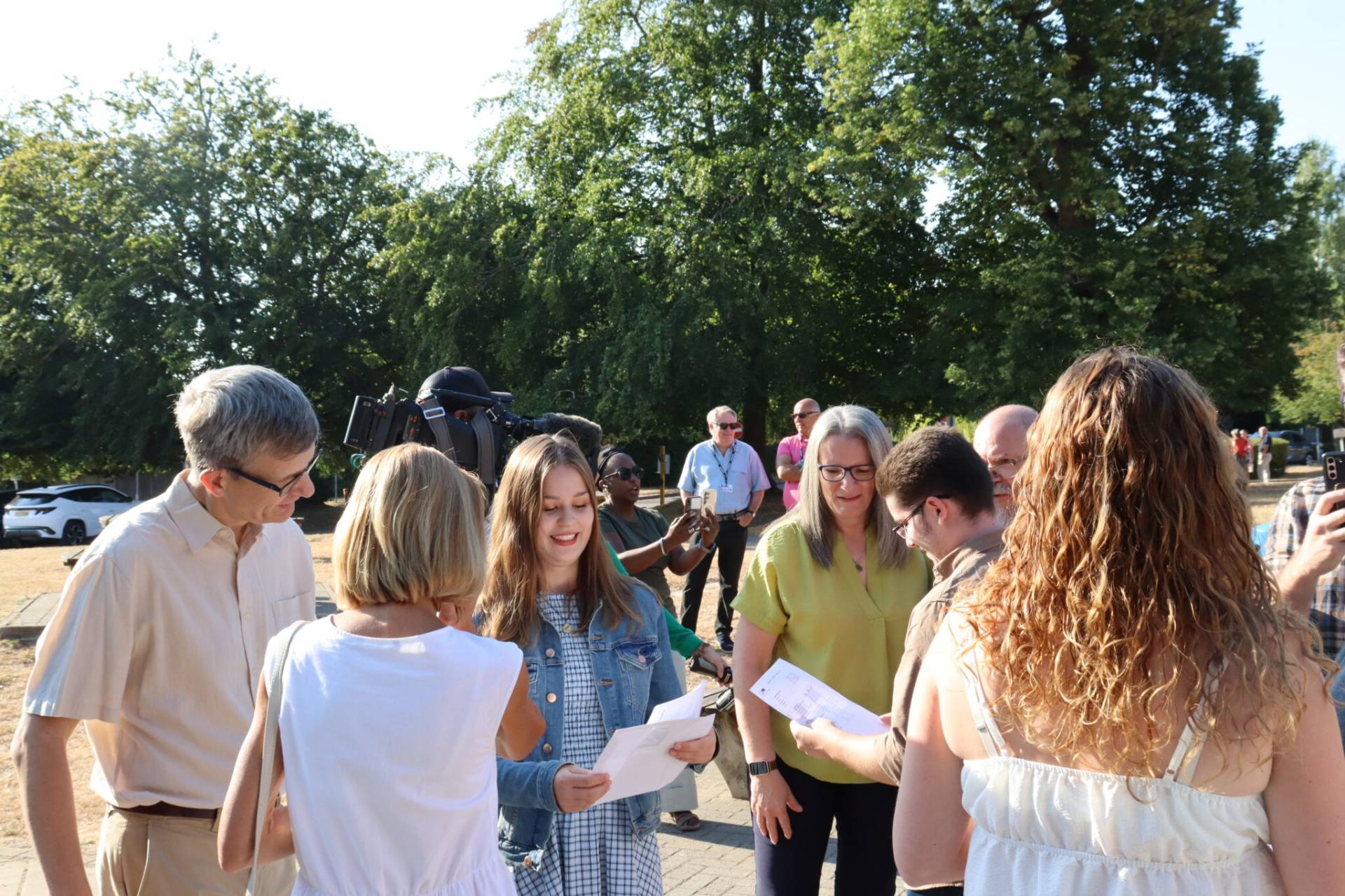 Group of people standing outside, looking at papers, with a camera crew behind them and tall green trees in the background.