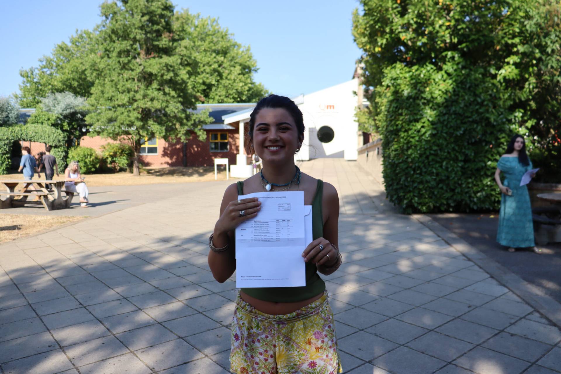 Smiling woman in a green top and floral skirt holds papers outdoors on a sunny campus with trees and a building in the background, facing the camera.