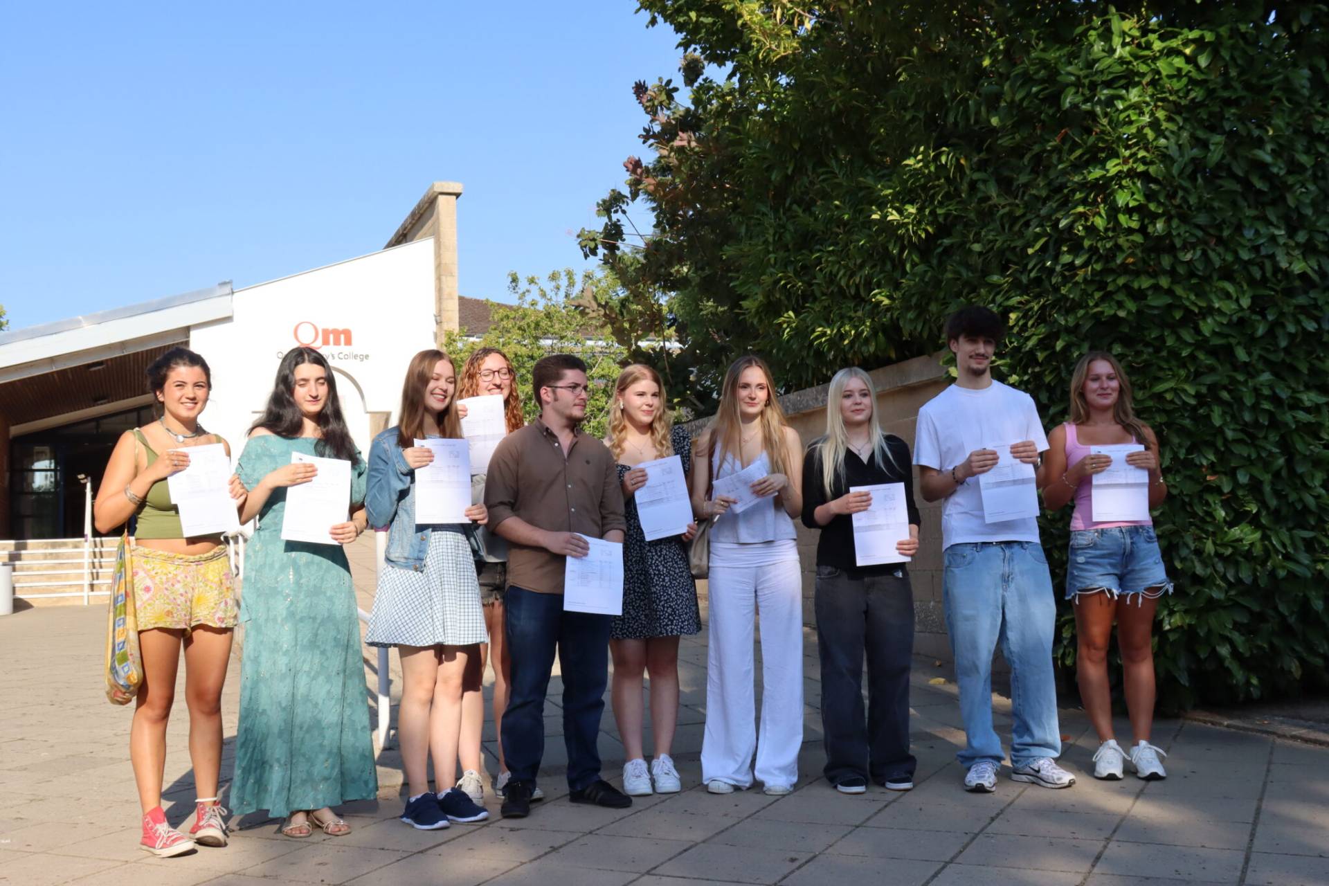 Group of young students holding certificates outdoors on a sunny day in front of a building and greenery.