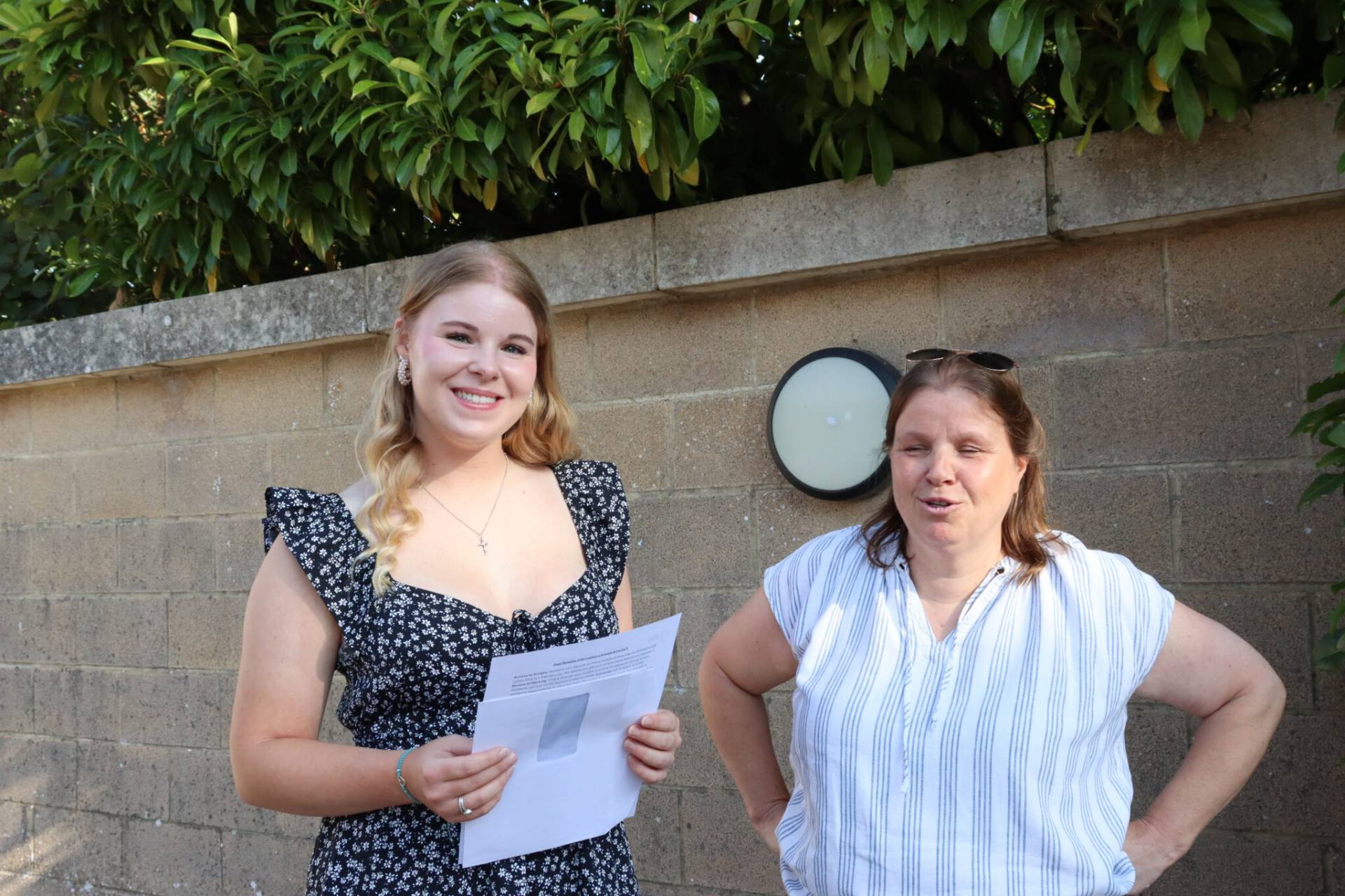 Two women posing outdoors in front of a concrete wall with greenery above; the left woman holds papers and smiles. The right woman wears a striped shirt and sunglasses on her head, smiling as well.