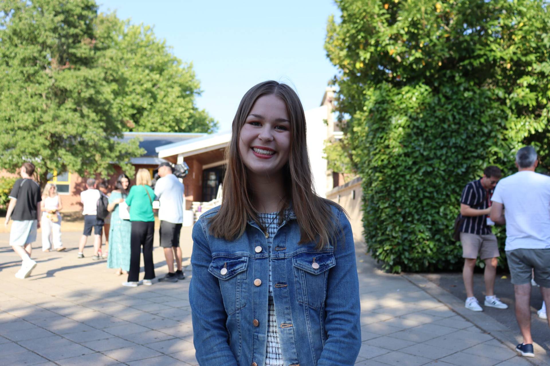 Young woman with a denim jacket smiling at the camera in an outdoor gathering with people in the background.
