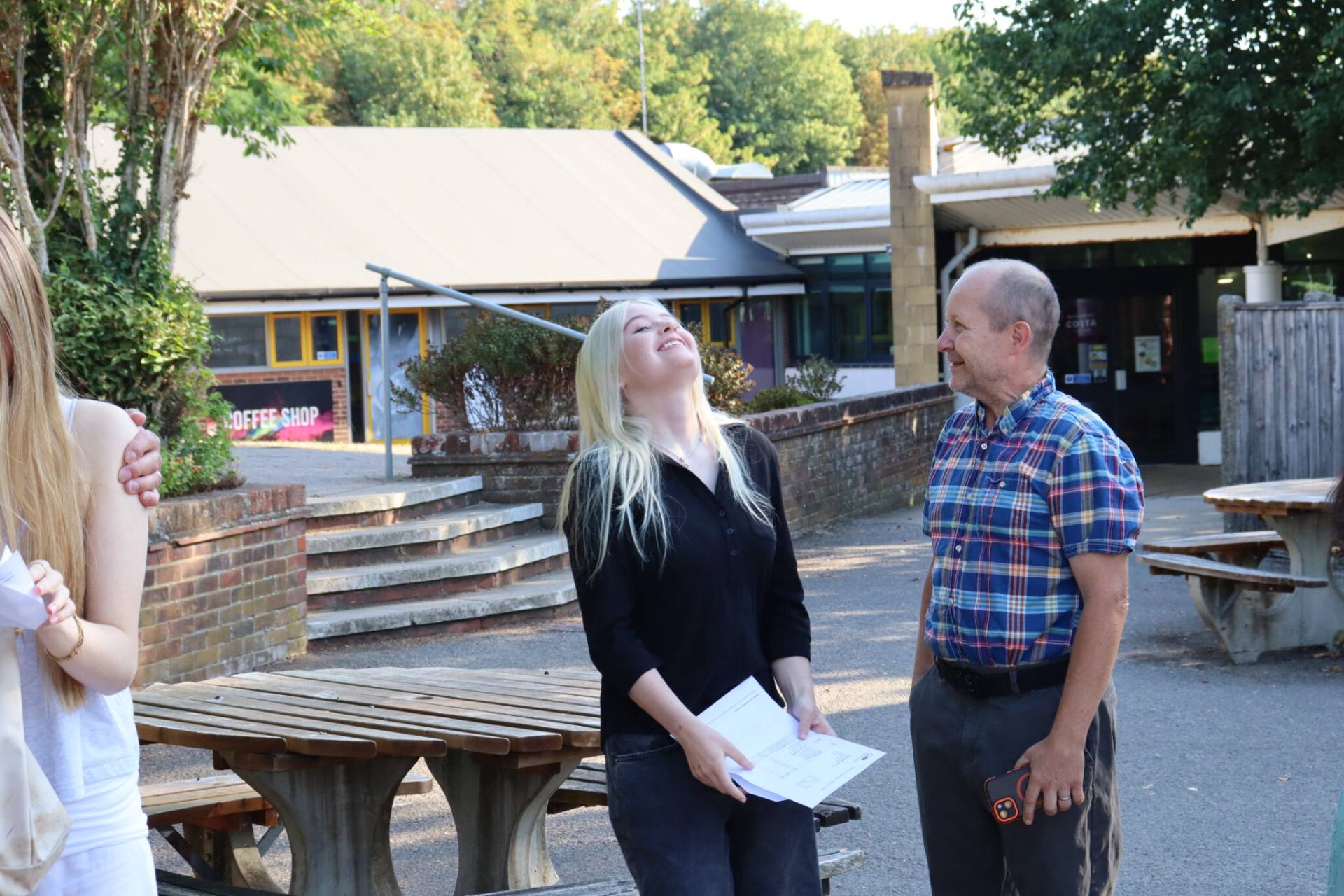 A smiling man in a plaid shirt and a young blonde woman stand outside, holding papers, near brick steps and wooden benches in a courtyard.