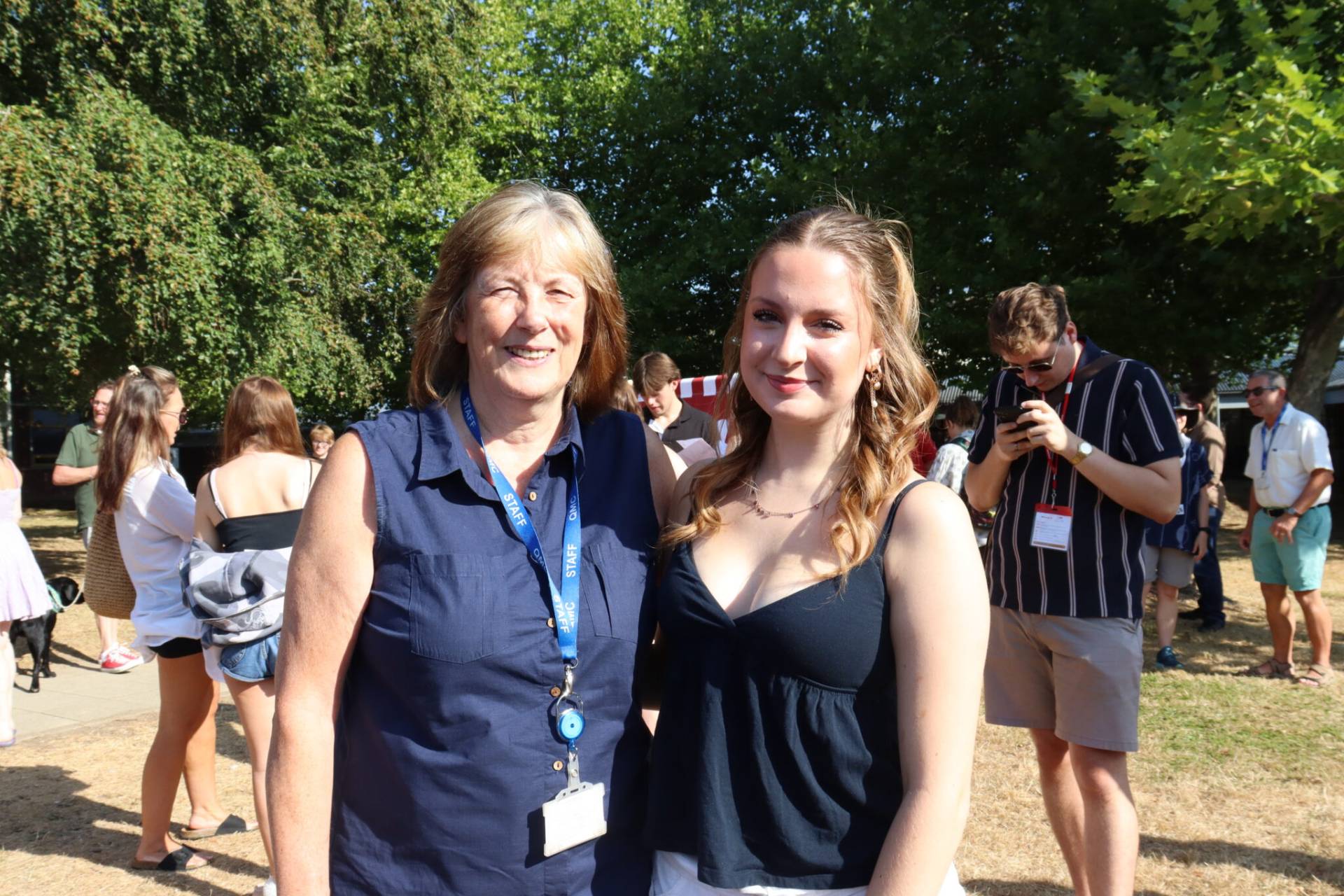 Two women pose for a photo at an outdoor event; an older woman in a blue sleeveless top with a staff badge stands beside a younger woman in a navy dress, with trees behind them.