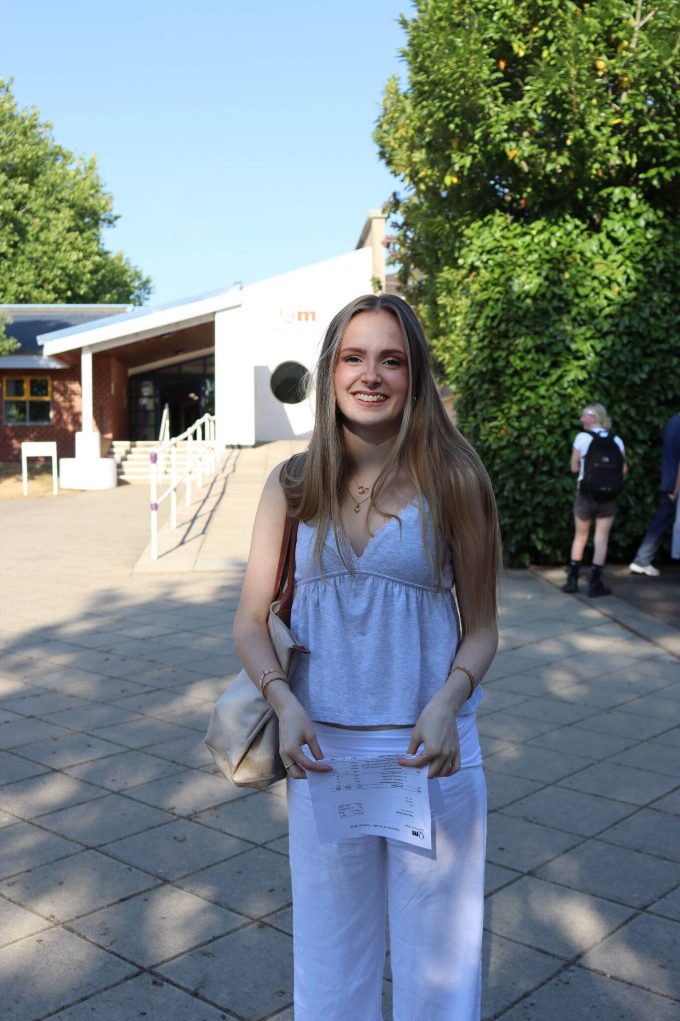 Smiling young woman standing on a paved square, holding a printed document and a beige bag.