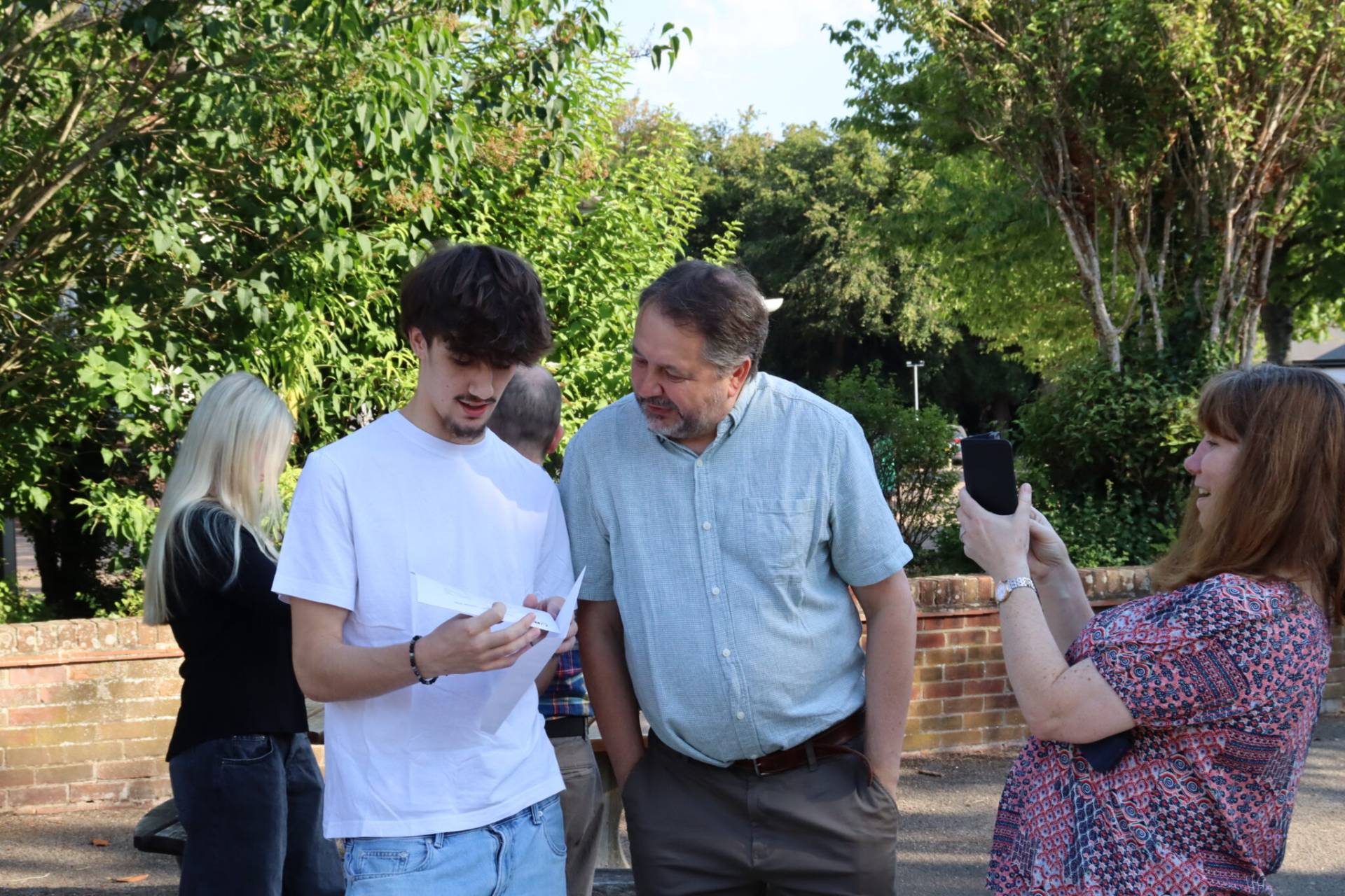 Several people stand outside around a brick wall, looking at papers together as one person photographs the moment with a smartphone.