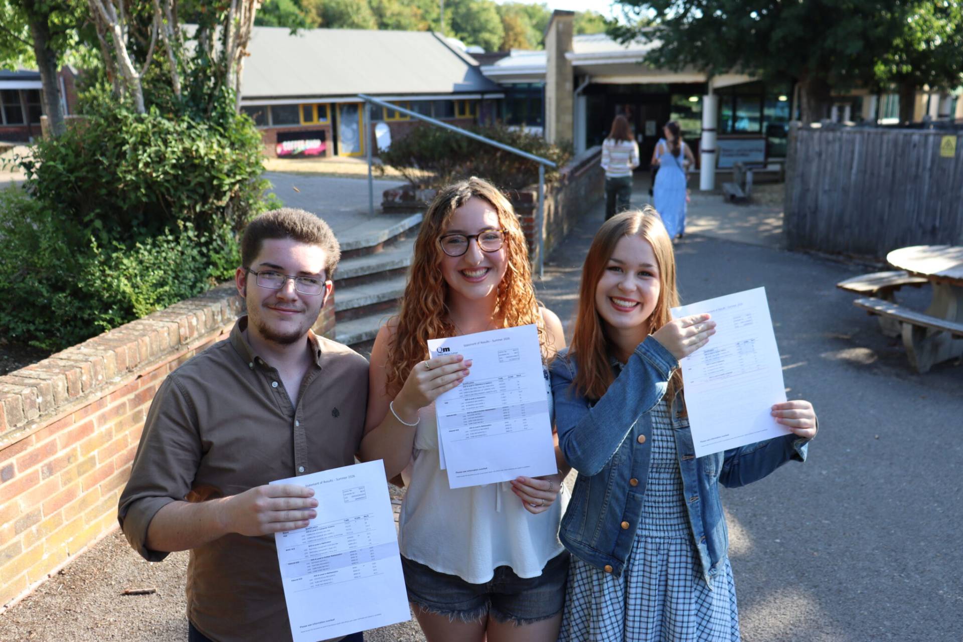 Three smiling young adults standing outdoors on a campus walkway, each holding a printed document or form. A brick wall and trees frame the left, with campus buildings in the background.