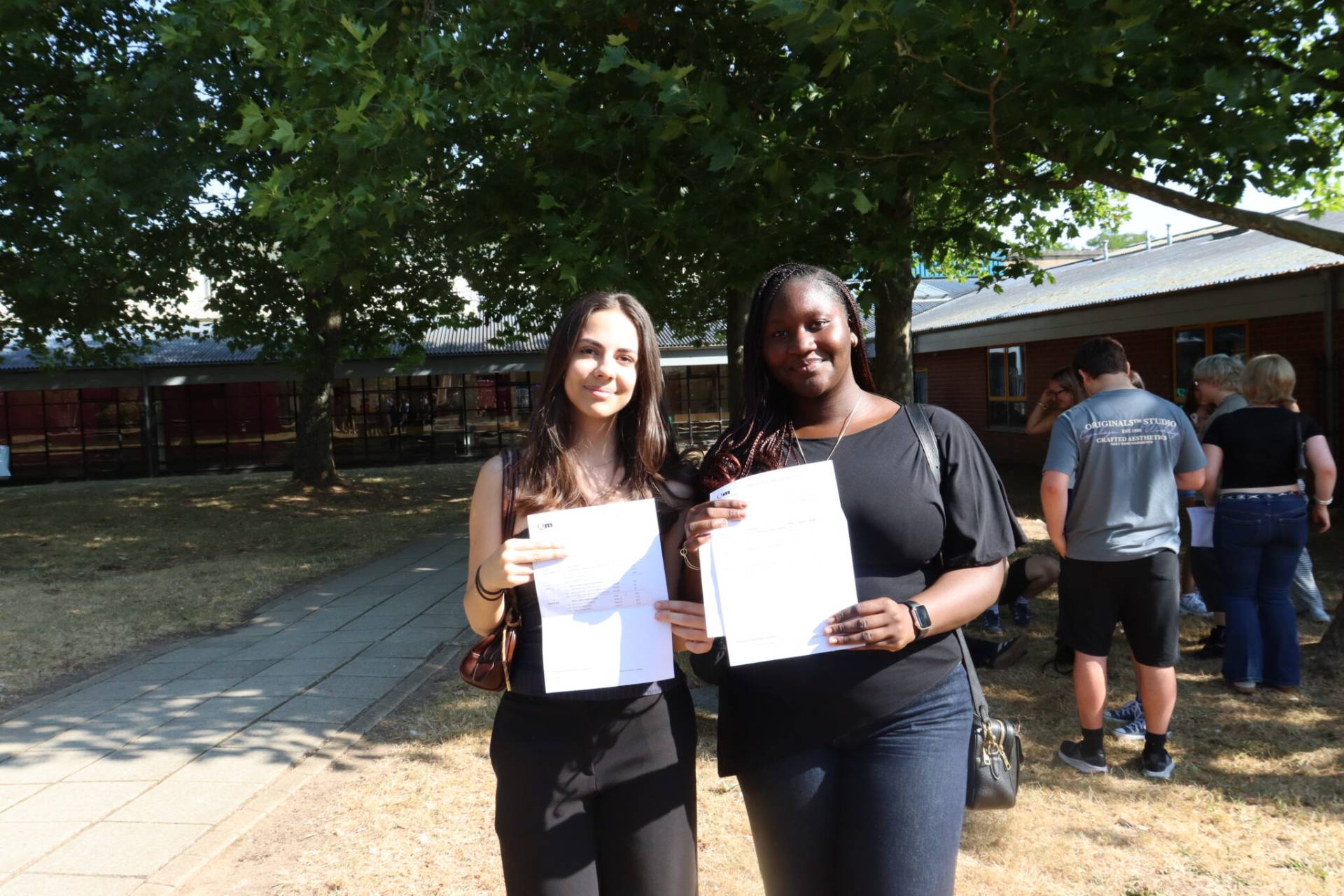 Two young women stand outdoors holding certificates and posing for a photo on a sunny campus walkway.