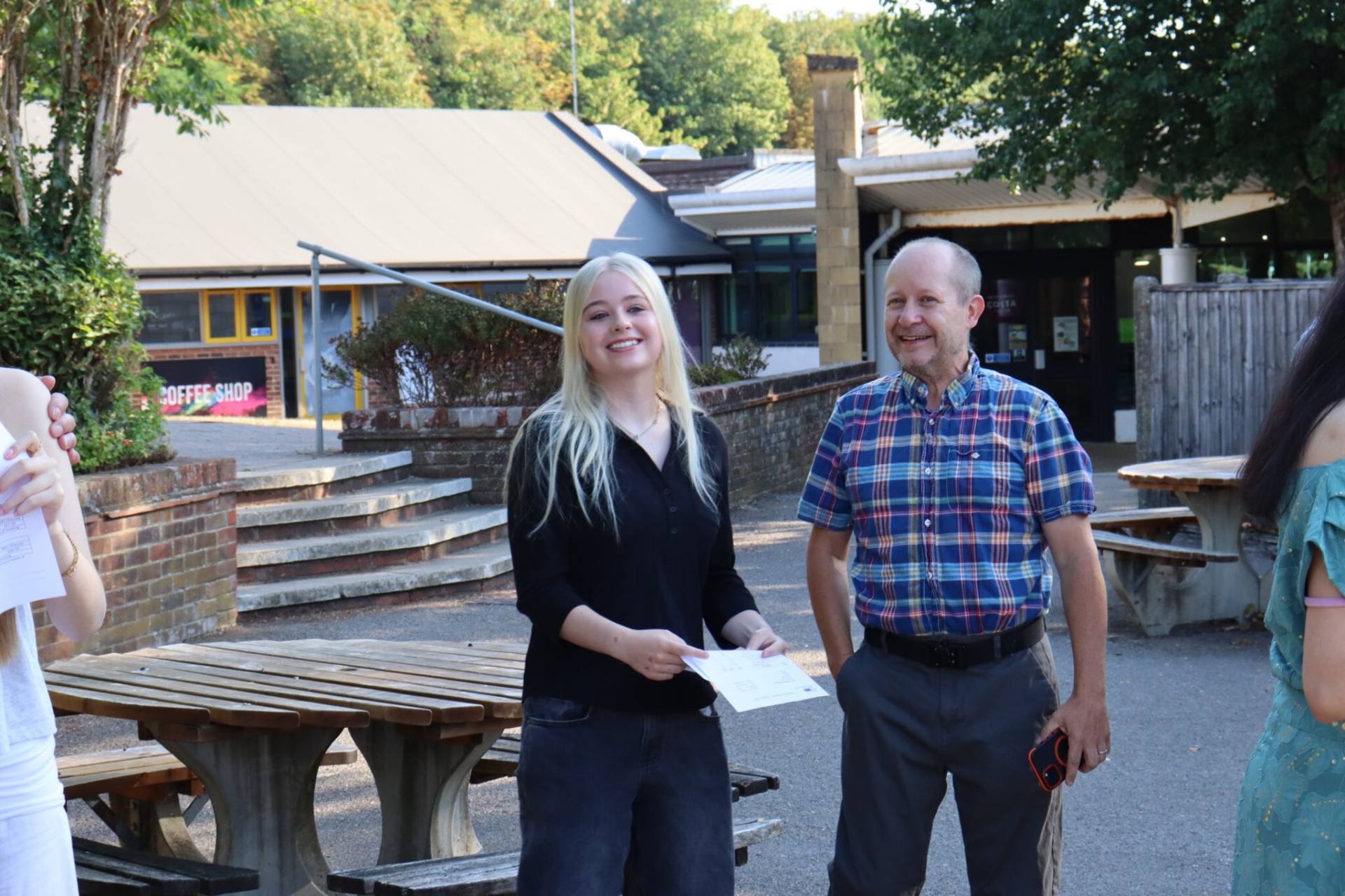 Smiling group of people outdoors near a wooden table, holding papers, in casual clothing.
