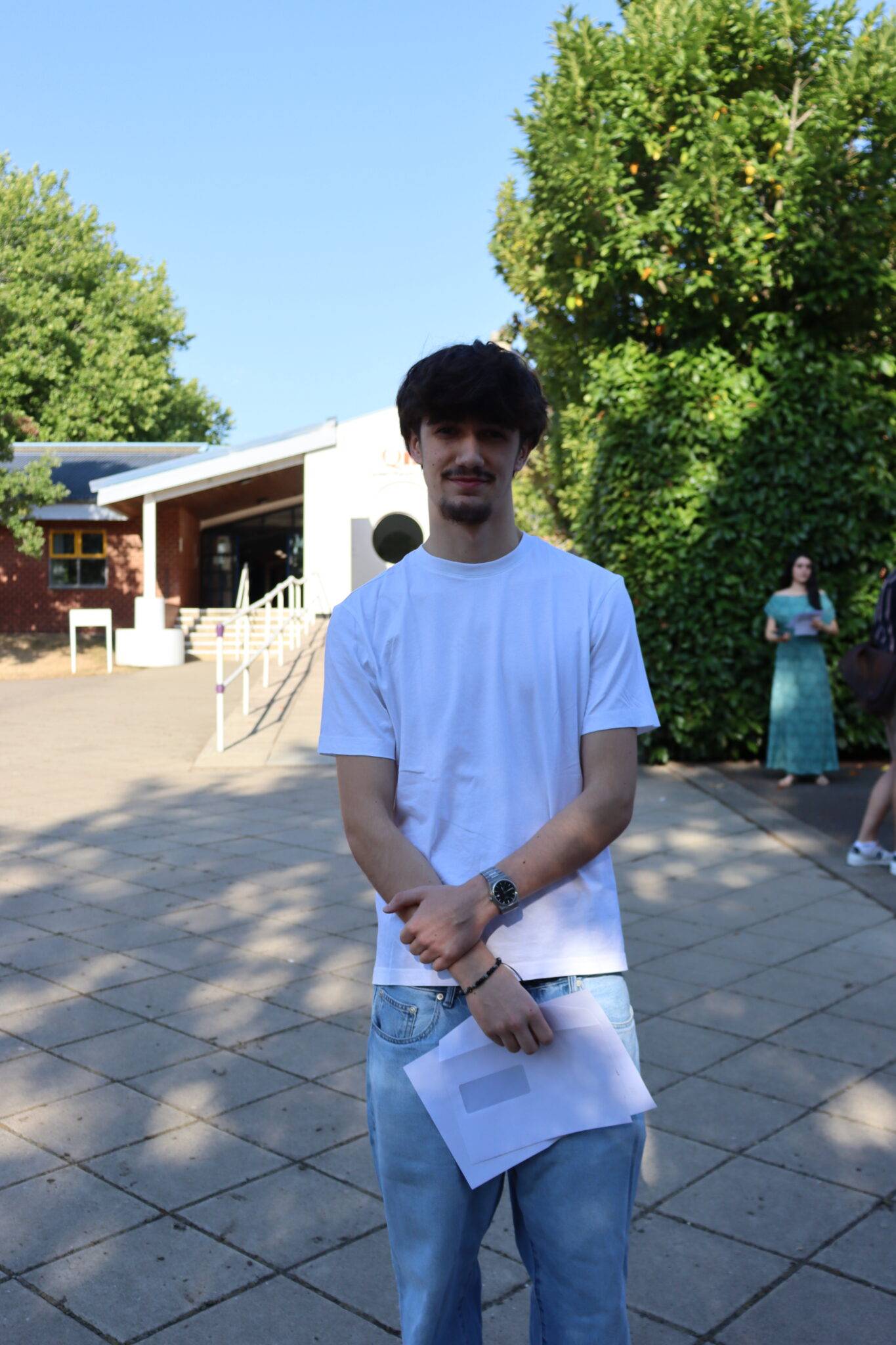 Young man in a white T-shirt and light jeans stands outdoors, holding papers; a woman in a green dress is in the background near bushes.