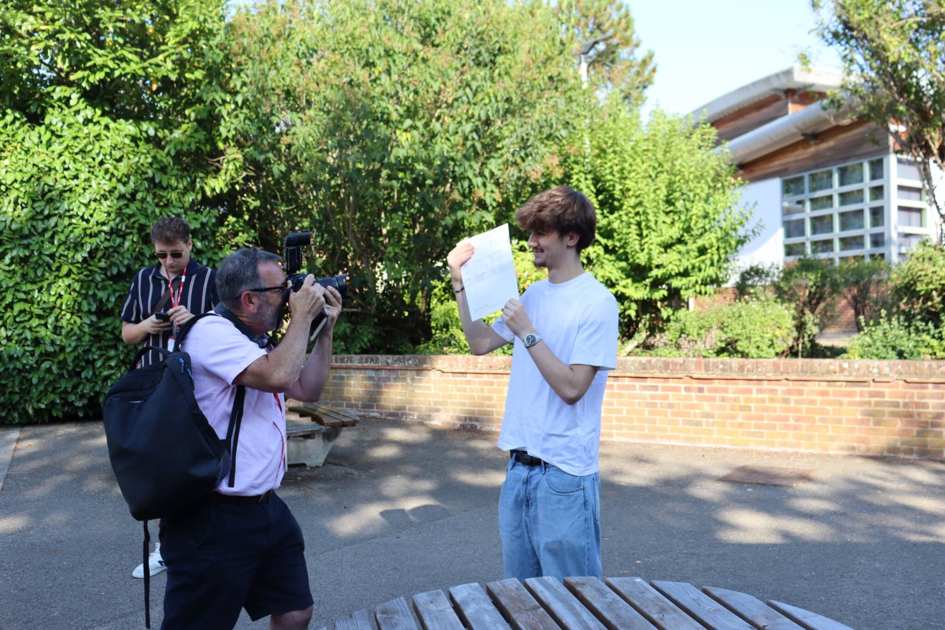 Photographer with camera aiming at a young man holding a sheet of paper outdoors, surrounded by greenery and a brick wall.