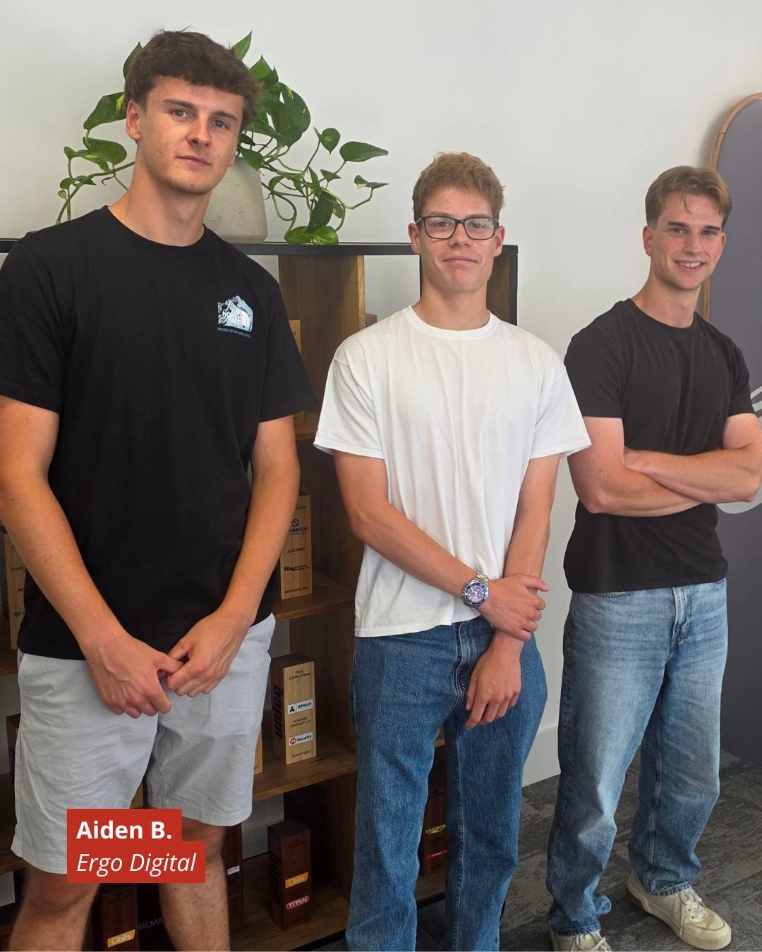 Three young men stand side by side in a casual office, left to right wearing black, white, and black tees; a plant and wooden shelves behind them; red badge shows 'Aiden B. Ergo Digital'.