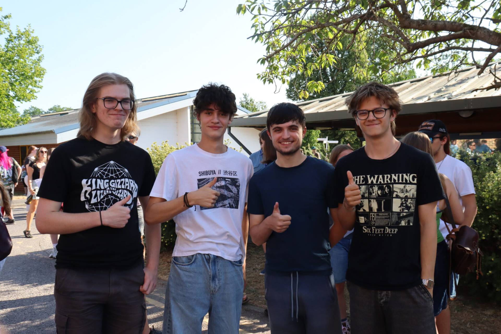 Four young men posing for a photo outdoors at a sunny gathering, giving thumbs up and smiling at the camera. They wear graphic T-shirts and casual jeans.