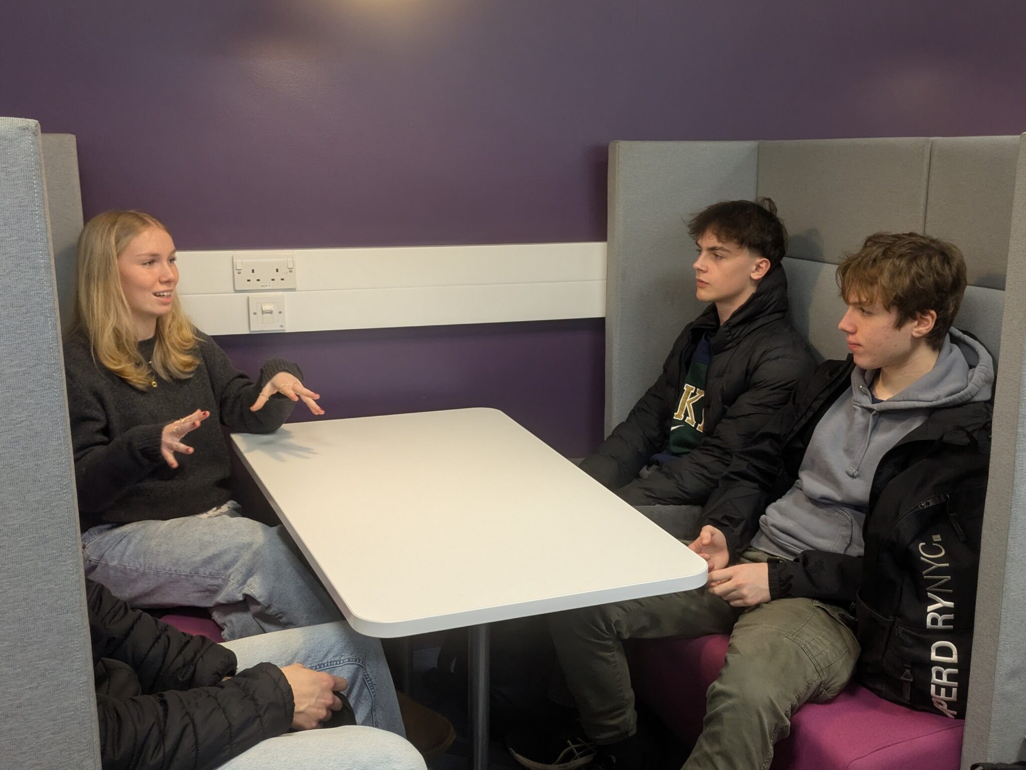 A group of young people are sitting around a white table in a booth with purple walls. One person is talking with their hands animatedly, while the other three listen.