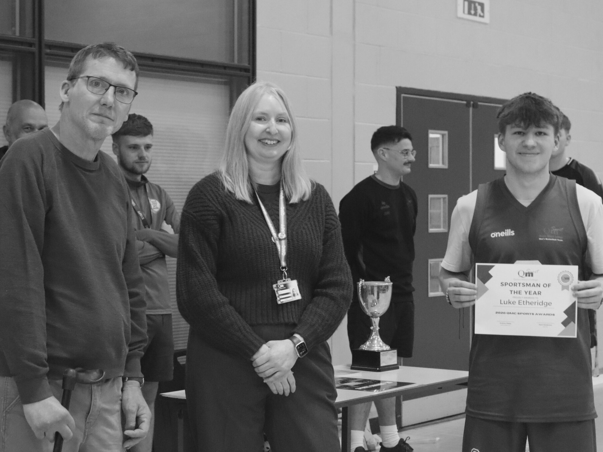 Group of people posing indoors for a photo at an awards event; a smiling woman in the center, a man in a dark shirt to her left, and a young man in a sports jersey on the right holding a certificate and trophy on a table nearby.