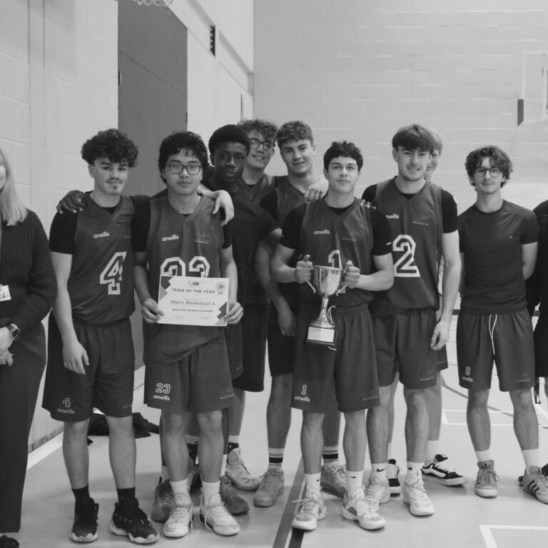 Group of basketball players in matching jerseys pose with a trophy and certificate in a gym, flanked by two adults.