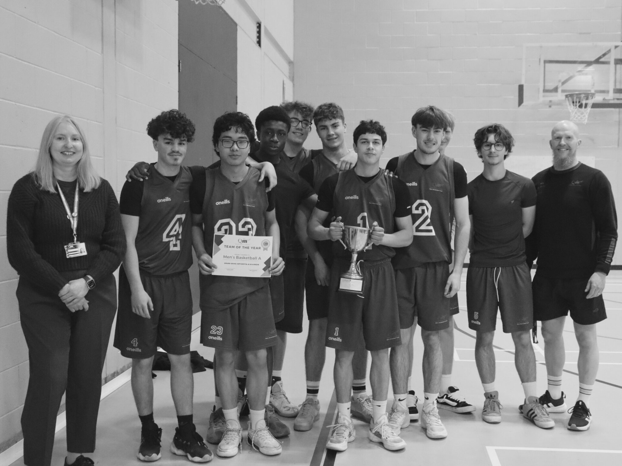 Group of basketball players in matching jerseys pose with a trophy and certificate in a gym, in between by two adults.