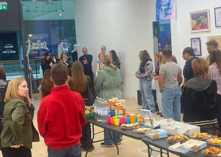 A group of people stand around tables laden with snacks and drinks at an indoor event, with artwork displayed on the walls.