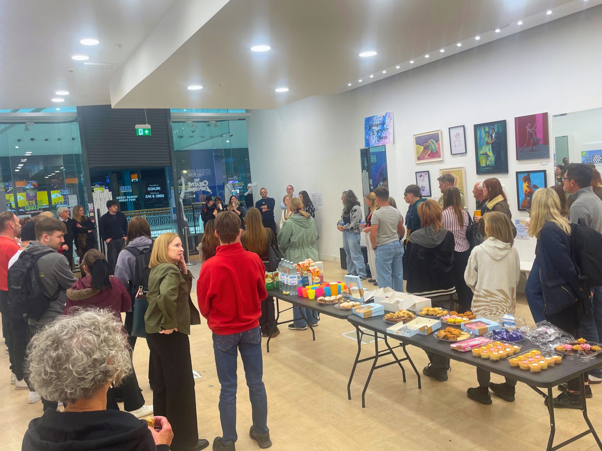A crowd of people gather in an art gallery, with paintings displayed on the walls and a table laden with snacks and drinks in the foreground.