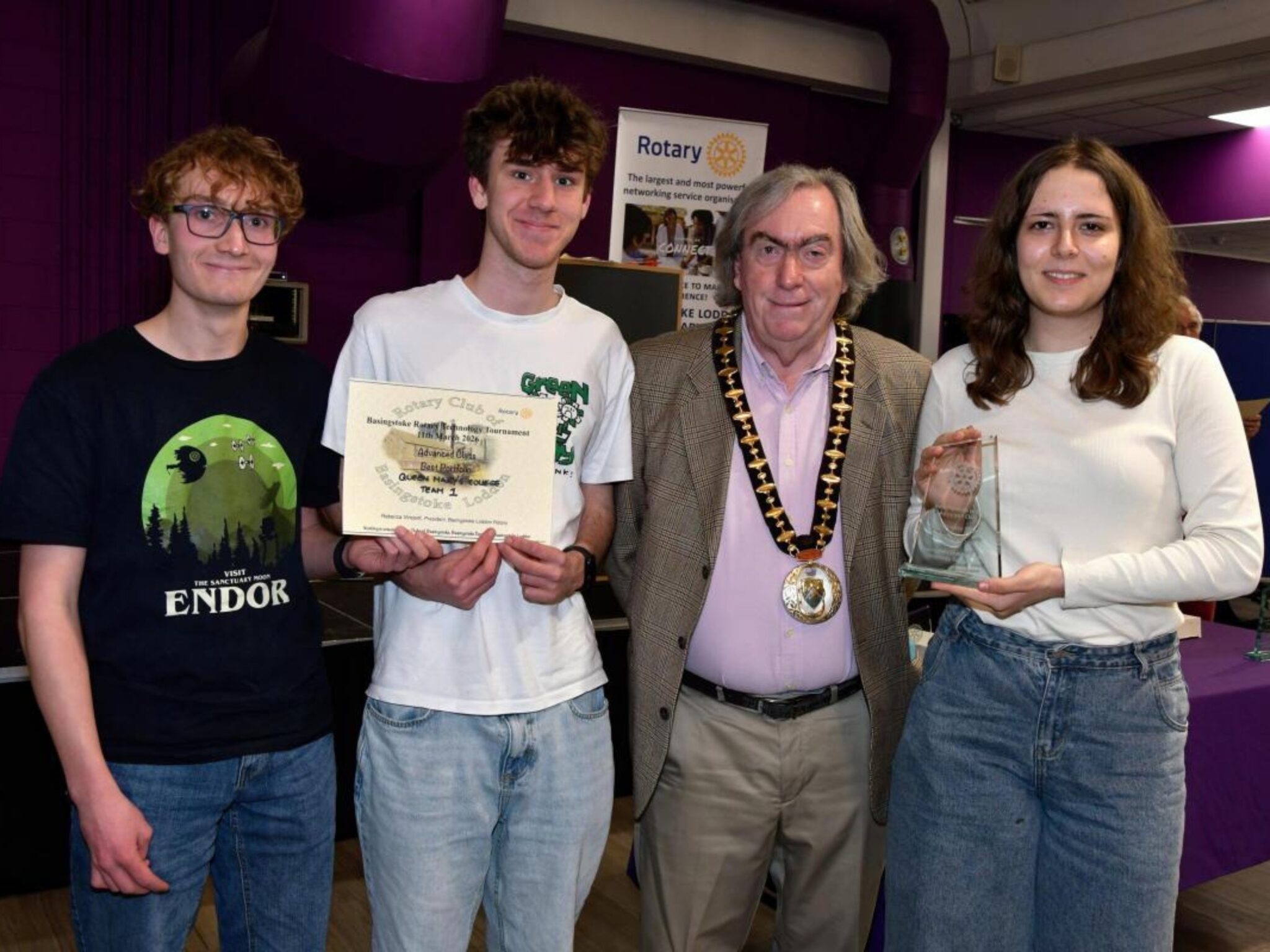 A man in ceremonial chains stands between three young people, two of whom are holding awards and certificates.