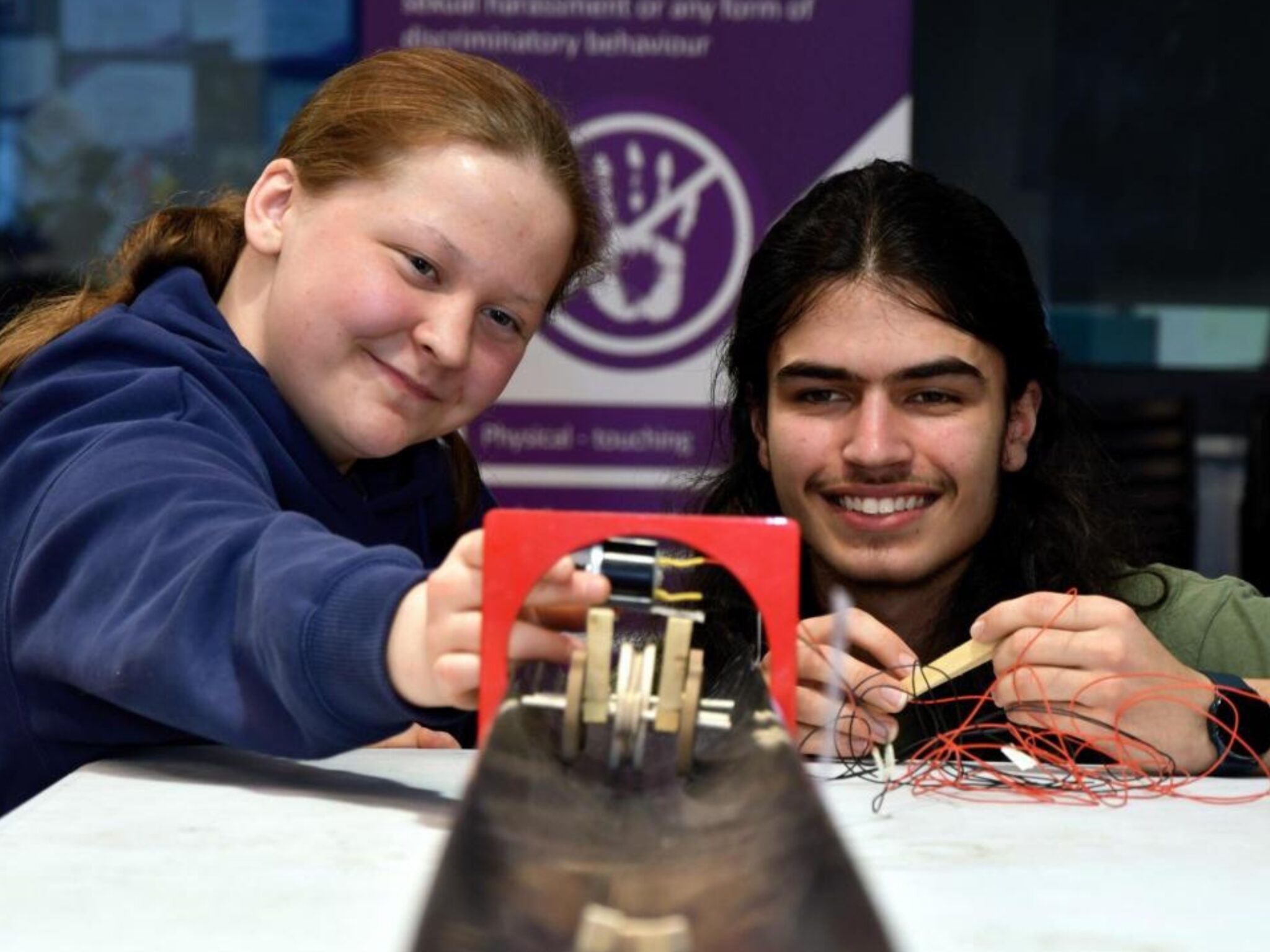 Two individuals are working on a project involving wires and a red contraption, smiling at the camera.