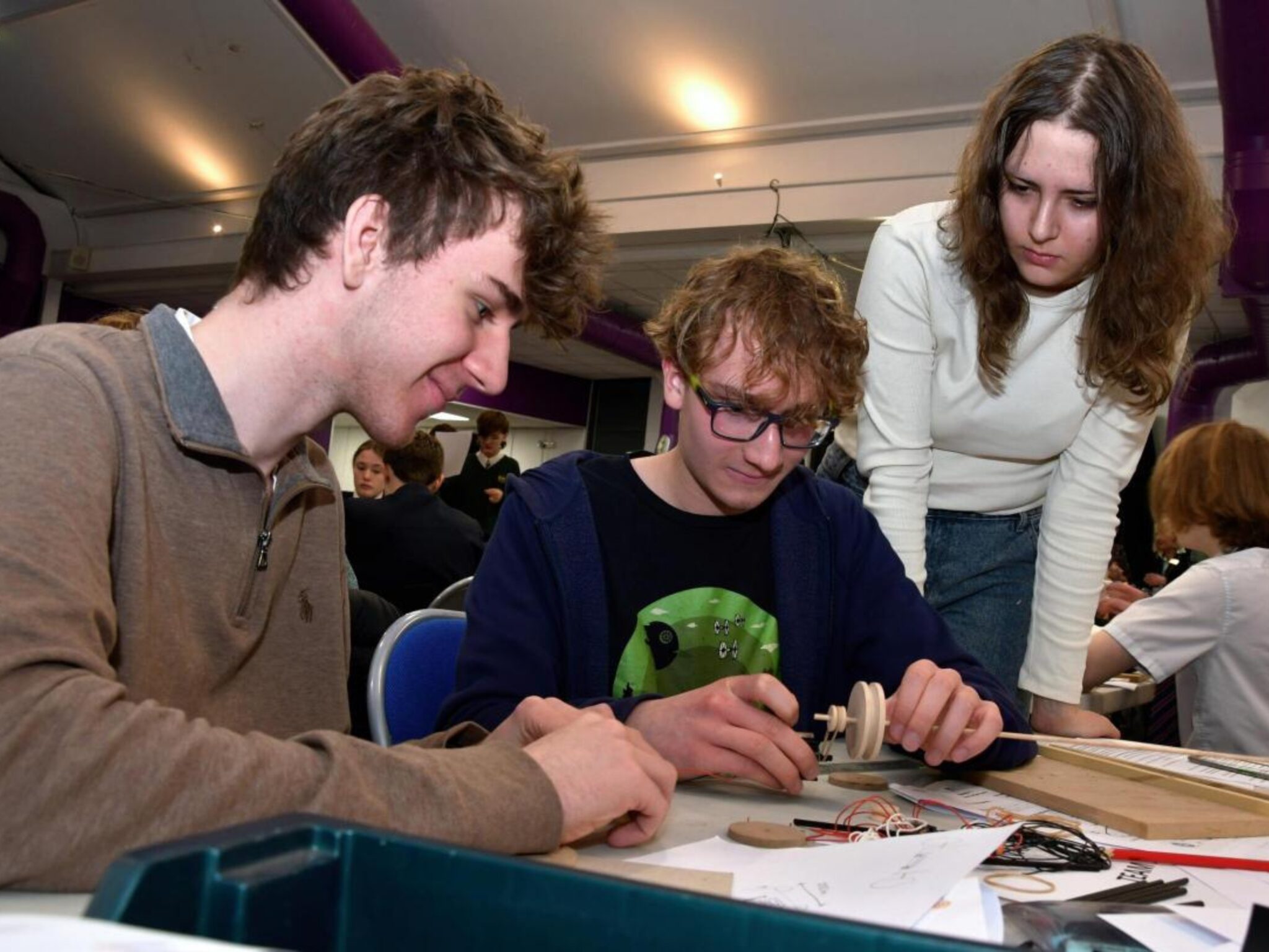 Three individuals are gathered around a table, engaged in a hands-on project involving wooden components and wires, in an engineering activity.