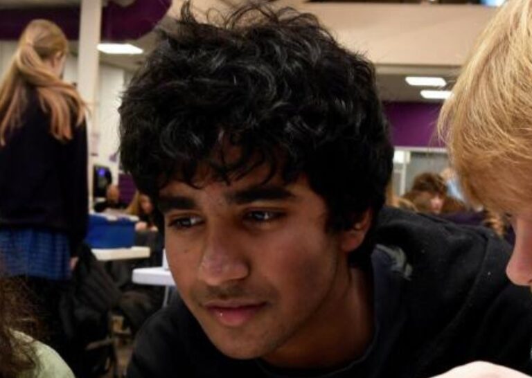 Three students sit together in a classroom, smiling as they look at something off-camera.