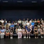 Large group of students sitting in an auditorium, posing for a class photo against dark blue walls.