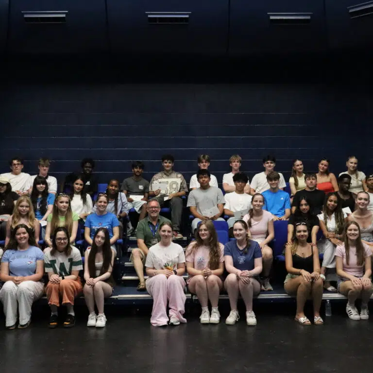 Large group of students sitting in an auditorium, posing for a class photo against dark blue walls.