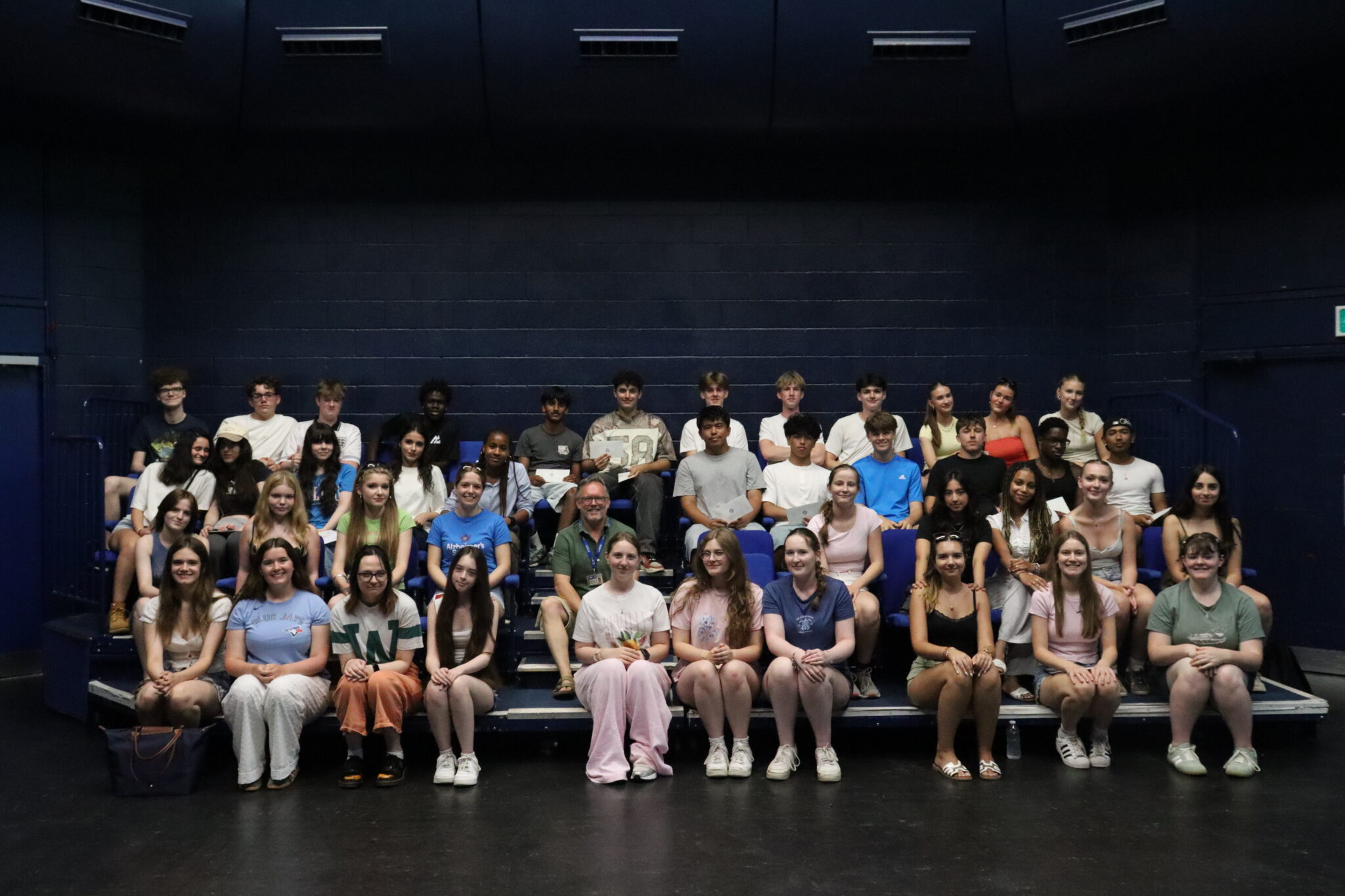 Large group of students sitting in an auditorium, posing for a class photo against dark blue walls.