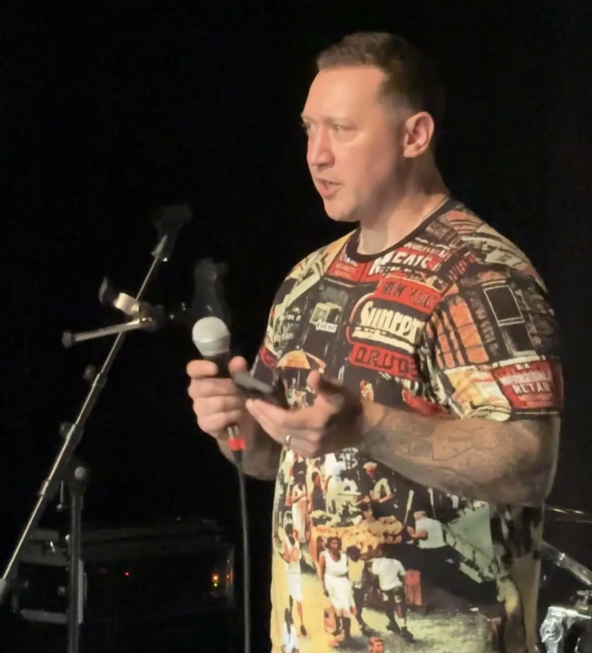 Man on stage speaking into a handheld microphone, wearing a busy graphic T‑shirt with collage images, dark background and stage gear nearby.