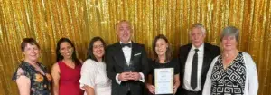 Seven adults in formal wear pose for a photo at an awards ceremony against a gold sequin backdrop; the man in the center holds a glass trophy and a certificate held by the woman beside him.