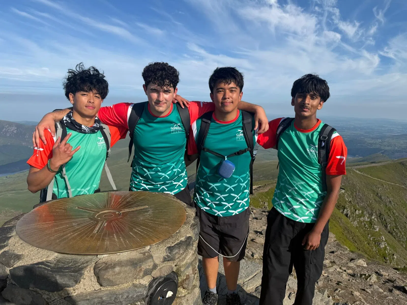 Four young hikers in teal shirts with backpacks pose on a mountain summit, arms around each other, with a circular bronze marker in the foreground and blue sky above.
