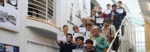 A group of students on a staircase proudly holding certificates at a campus awards event.