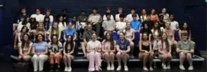 Large group of students seated on blue gym bleachers for a class photo in a gymnasium.