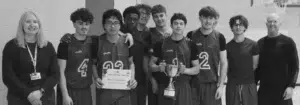 Group of teammates in basketball uniforms posing with a coach and a woman in a gym; one player holds a certificate and another holds a trophy, all smiling after a win.