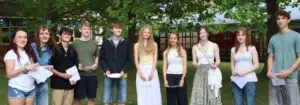 Ten teenagers stand in a row on a grassy campus lawn, holding papers and smiling at the camera.