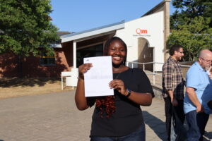 Smiling woman holding a document outside Queen Mary’s College, with the college sign visible above the entrance.