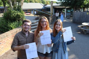 Three smiling young adults standing outdoors on a campus walkway, each holding a printed document or form. A brick wall and trees frame the left, with campus buildings in the background.