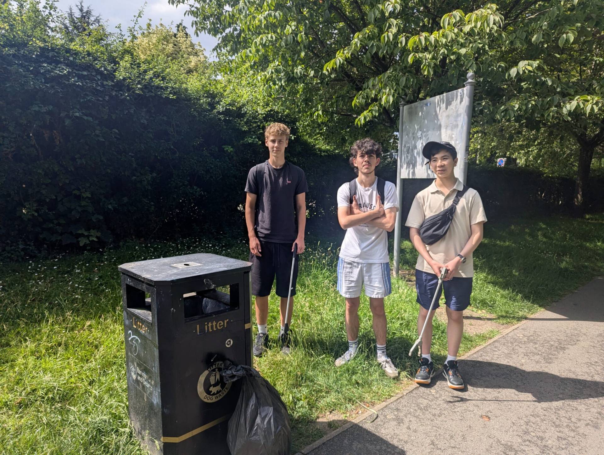 Three young men stand by a black litter bin with tied trash bags in a sunny, grassy park.